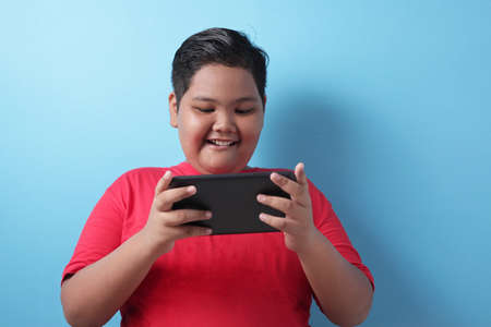 Portrait of fat Asian boy plays online game on his tablet, smiling happy face, kid with gadget...