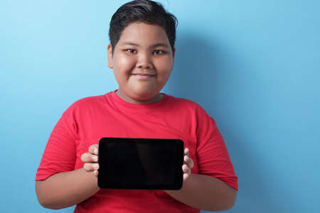 Happy smiling fat Asian boy shows his tablet screen, tablet smart phone mock up, against blue backgroundの写真素材