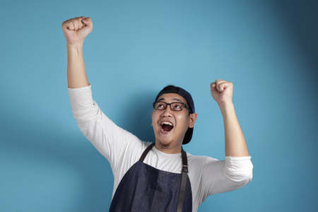 Portrait of happy excited proud Asian chef or waiter dancing happily espressing victory, against blue backgroundの写真素材