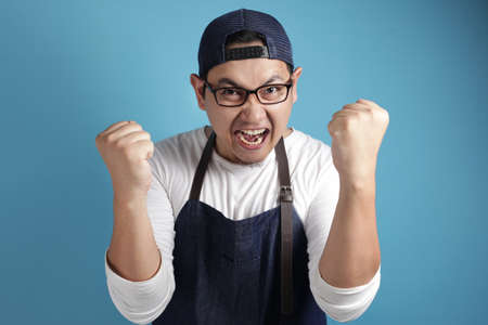 Portrait of happy excited proud Asian chef or waiter shows winning gesture, against blue backgroundの写真素材