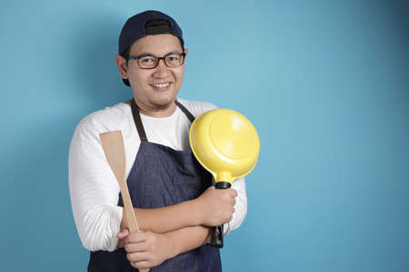 Portrait of Asian male chef smiling happy while holding kitchen tools, chef holding spatula and pan,...