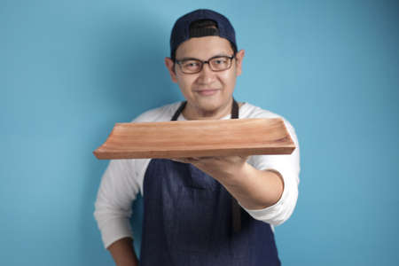 Portrait of Asian male chef or waiter looks happy and proud presenting something on his empty wooden plate, copy space meal menu conceptの写真素材