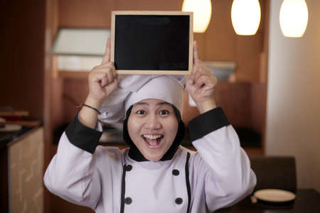 Portrait of Asian female chef looks happy and proud showing empty blackboard, presenting blank writing template, copy space meal menu conceptの写真素材