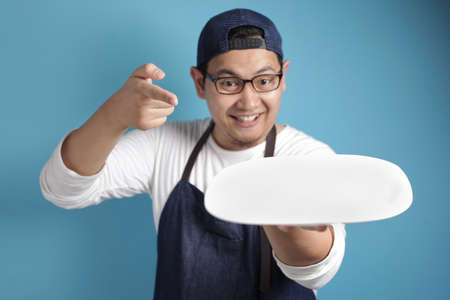 Portrait of Asian male chef or waiter looks happy and proud presenting something on his empty white plate, copy space meal menu conceptの写真素材