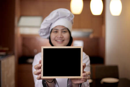 Portrait of Asian female chef looks happy and proud showing empty blackboard, presenting blank writing template, copy space meal menu conceptの写真素材