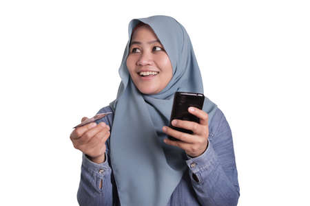 Portrait of Asian muslim woman smiling and making purchase buying online from her mobile phone,...
