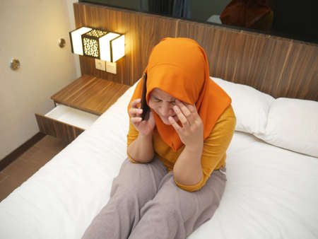 Portrait of young Asian muslim woman crying sad when looking at her phone, worried expression. Receiving bad news on phone conceptの写真素材