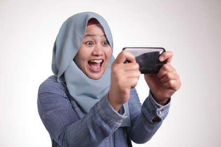 Happy Asian muslim woman excited to play games on her smart phone, isolated on whiteの写真素材