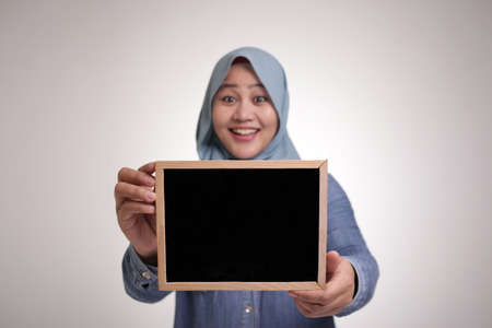 Portrait of smart happy successful Asian muslim woman wearing hijab smiling at camera while holding and showing empty blackboard or chalk board with copy spaceの写真素材