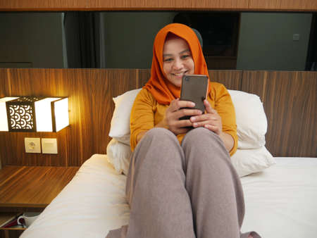 Happy Asian muslim woman wearing hijab smiling when reading text message or chat on her phone while...