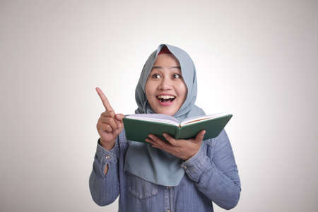 Portrait of young Asian muslim woman pointing finger up and smiling having good idea gesture while...