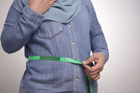 Portrait of funny fat Asian muslim woman shocked worried to see her big belly, measuring waist,...