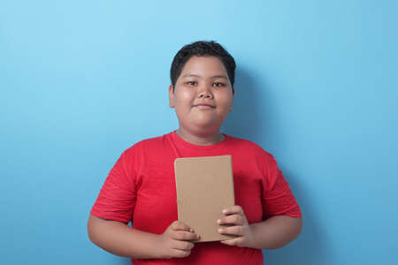 Asian boy student smiling happily looking at the camera while holding books, educational and knowledge concepts, in front of a blue backgroundの写真素材