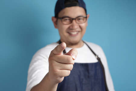 Portrait of young male Asian chef or waiter smiling and pointing forward, looking at camera. Choosing someone concept with selective focus, against blue backgroundの写真素材