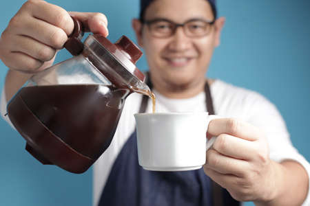 Portrait of Asian male chef or waiter smiling while pouring coffee to a cup, offering coffee concept, against blue backgroundの写真素材