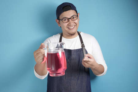 Portrait of Asian male chef or waiter smiling while offering fresh red syrup beverage, offering cocktail concept, against blue backgroundの写真素材