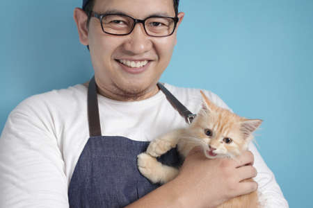 Happy Asian man smiling while holding his kitten. Animal lover with his cat pet concept, against blue backgroundの写真素材