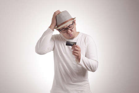 Portrait of attractive unhappy Asian man holding a credit card with sad, confused and shocked expression, against white wall. The concept of a limit of money and bankruptcyの写真素材