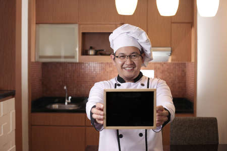Portrait of Asian male chef looks happy and proud showing empty blackboard, presenting blank writing template, copy space meal menu conceptの写真素材