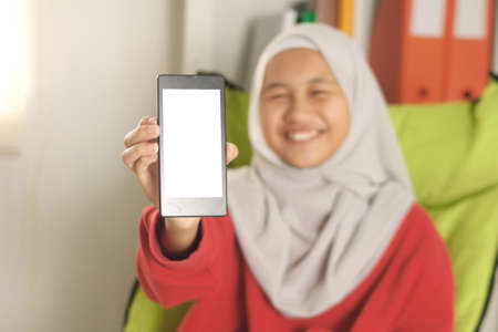 Happy cheerful Asian muslim girl wearing hijab shows her smart phone, phone template mock up, selective focus on phoneの写真素材