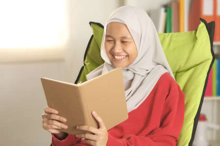 Happy Asian muslim girl wearing hijab smiling while reading book, leisure activity casual lifestyle, Islamic religion ethnicityの写真素材