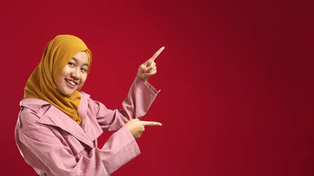 Portrait of attractive beautiful muslim teenage girl wearing hijab  smiling and pointing to the side with copy space, presenting something, marketing concept against red backgroundの写真素材