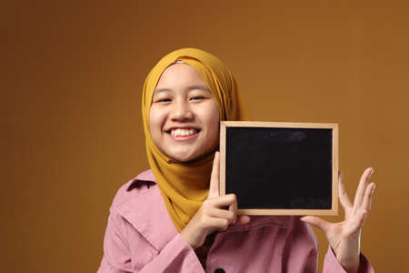 Portrait of smart young cute teenage Asian muslim girl wearing hijab smiling at camera while holding and showing empty blackboard or chalk board against yellow background, copy spaceの写真素材
