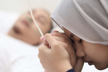 Asian muslim girl worried of her father being sick in hospital, she holds her fathers hand and pray for his health, family health insurance conceptの写真素材