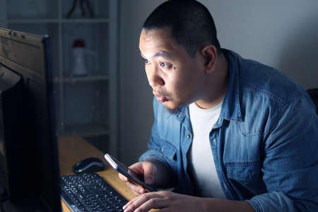 Young Asian man using smart phone and computer until late night, he is working or studying hard to...