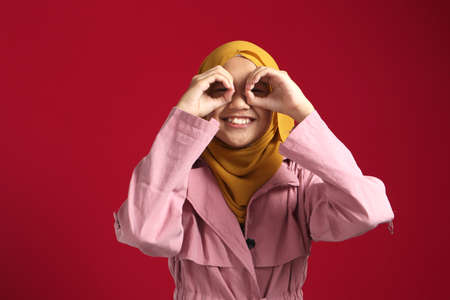 Happy young beautiful Asian muslim teenage girl making binoculars hands looking at camera and smiling, studio shot on red backgroundの写真素材