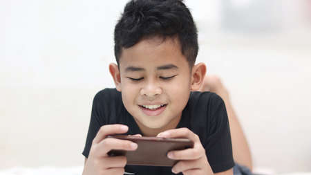 Asian boy lying on bed playing games on his phone, spending too much time using smart phone, gadget...