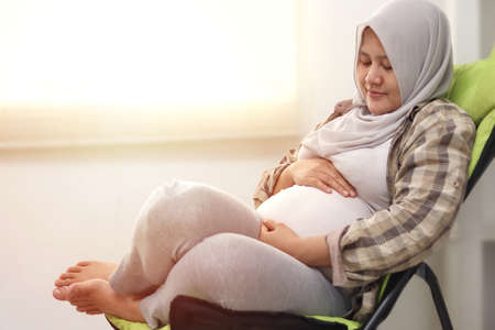 Beautiful pregnant Asian muslim woman relaxing on deck chair in the afternoon, enjoys her time while holding her bellyの写真素材