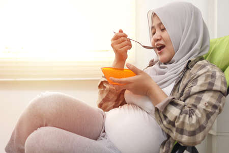 Happy Asian muslim pregnant mom sitting on deck chair and eating her healthy food for her baby in...