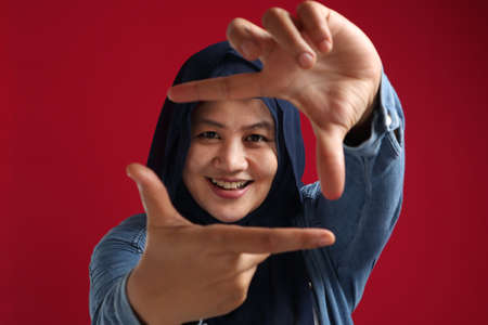 Smiling young Asian muslim woman wearing hijab making frame with fingers, red backgroundの写真素材