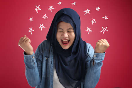 Portrait of success beautiful muslim businesswoman wearing hijab screaming shouting and shows winning victory gesture over red backgroundの写真素材