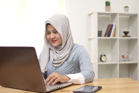 Happy young Asian muslim woman freelancer wearing hijab working on computer at home. Attractive...