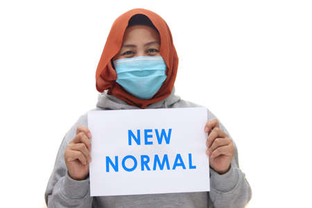 Asian muslim woman wearing hijab and facial mask shows note paper with new normal message written on...