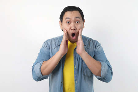 Portrait of young funny Asian man in white shirt, gesturing shocked or surprised expression with...