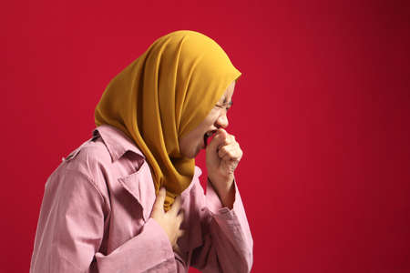 Asian muslim teenage girl covering her mouth when coughing; against red background