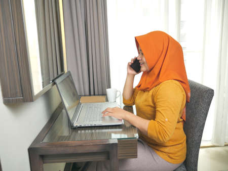 Asian muslim woman wearing hijab smiling while talking on phone, Successful businesswoman entrepreneur working at home. Wireless communication or work from home conceptの写真素材