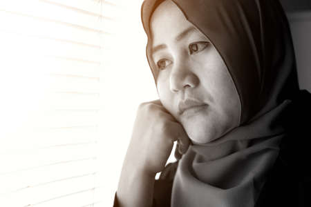 Close up portrait of sad Asian muslim woman looking out of window, lonely sadness conceptの写真素材
