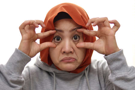 Tired muslim woman open her eyes with finger, trying to keep awake, close up portrait isolated on whiteの写真素材
