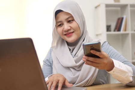 Asian muslim woman work in office, using phone and laptop, happy smiling facial expression, online...