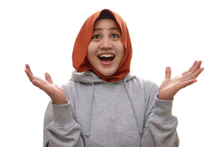 Happy Asian muslim woman shows happy surprised winning gesture greeting expecting something from above, isolated on whiteの写真素材
