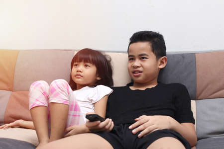 Young Asian boy and girl sibling sitting on the couch and watching tv, indoor home activity when school holidaysの写真素材