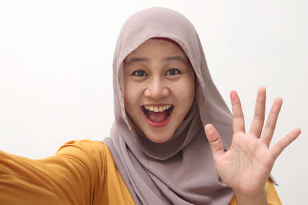 Happy cheerful beautiful Asian muslim lady wearing hijab waving to camera, doing selfie or video...