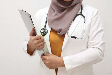 Asian muslim female doctor wearing hijab with stethoscope in white coat holding clipboard, ready to serve her medical patient, write medical record diagnosisの写真素材