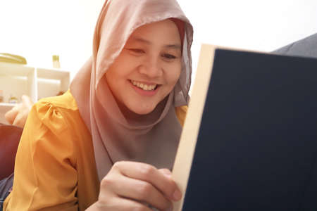 Beautiful Asian muslim woman reading book while sitting on sofa, girl enjoys her time by doing leisure activity, happy cheerful expressionの写真素材