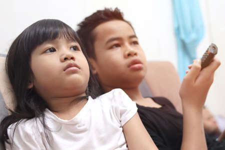 Young Asian boy and girl sibling sitting on the couch and watching tv, indoor home activity when school holidaysの写真素材
