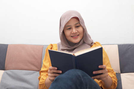 Beautiful Asian muslim woman reading book while sitting on sofa, girl enjoys her time by doing leisure activity, happy cheerful expressionの写真素材